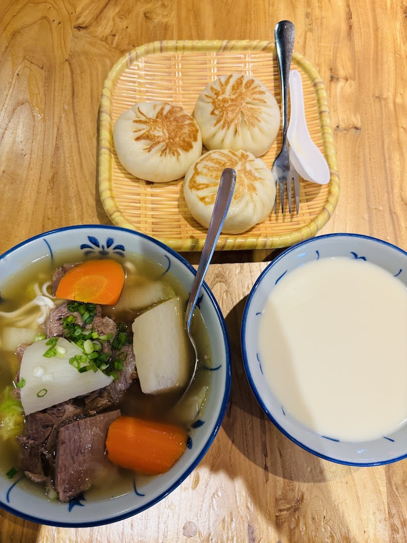 A delicious meal featuring beef noodle soup, pan-fried buns, and a bowl of soy milk on a wooden table.