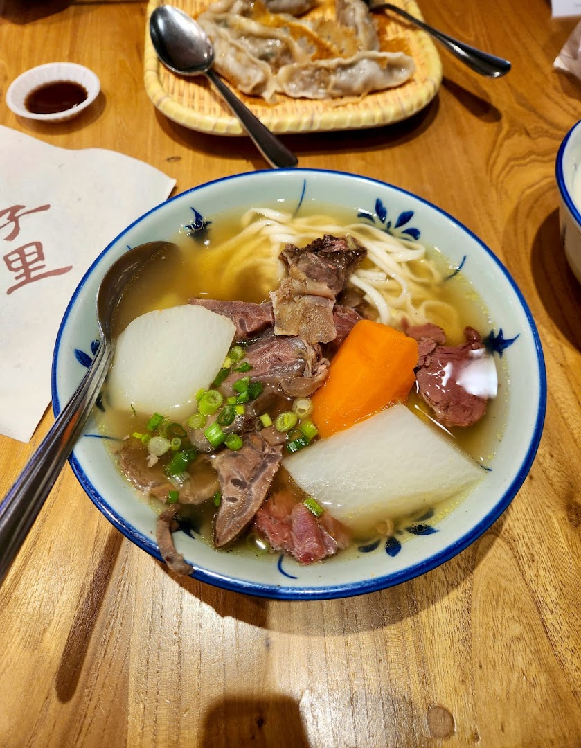A hearty bowl of beef noodle soup with vegetables and a side of dumplings on a wooden table.