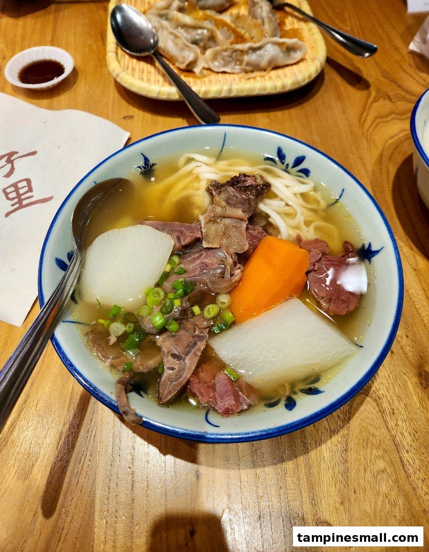 A hearty bowl of beef noodle soup with vegetables and a side of dumplings on a wooden table.