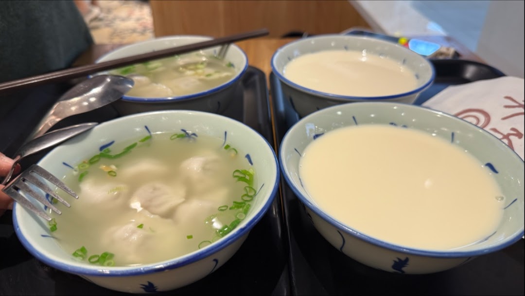 Four bowls of food and drinks, including dumplings/wontons and soy milk, served on trays.