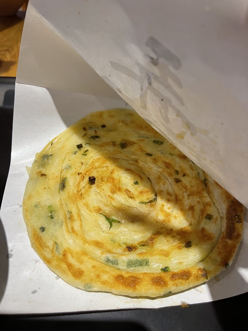 A close-up of a freshly cooked, golden-brown scallion pancake served in white paper packaging.