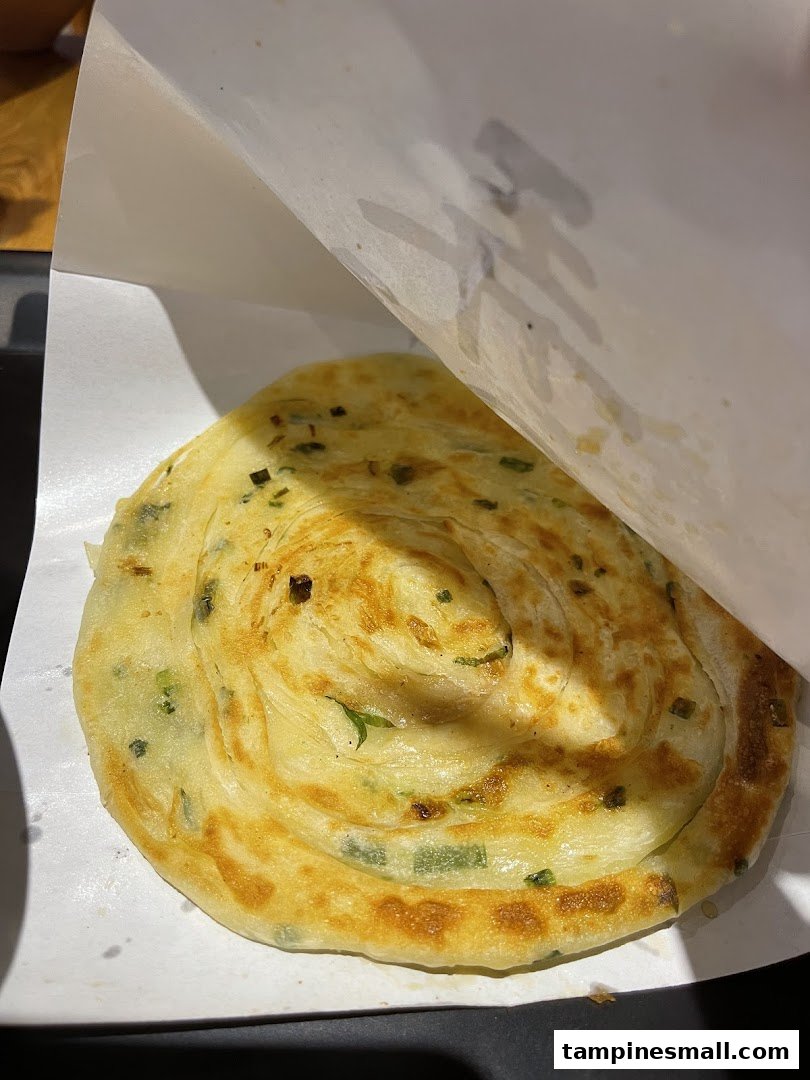 A close-up of a freshly cooked, golden-brown scallion pancake served in white paper packaging.