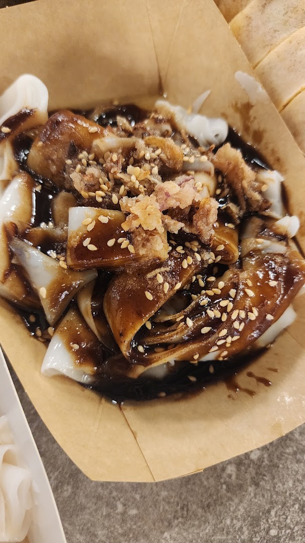 Close-up of savory rice rolls (Chee Cheong Fun) with dark sauce, sesame seeds, and crispy toppings in a paper bowl.