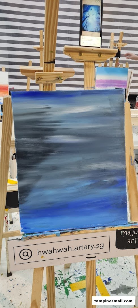 A blue and grey abstract painting on an easel, with a phone displaying a reference image.