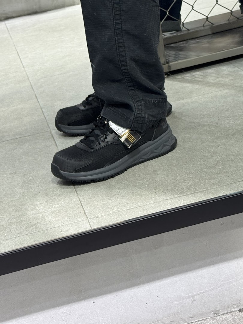A person trying on a pair of black athletic shoes with a tag visible, reflected in a mirror.
