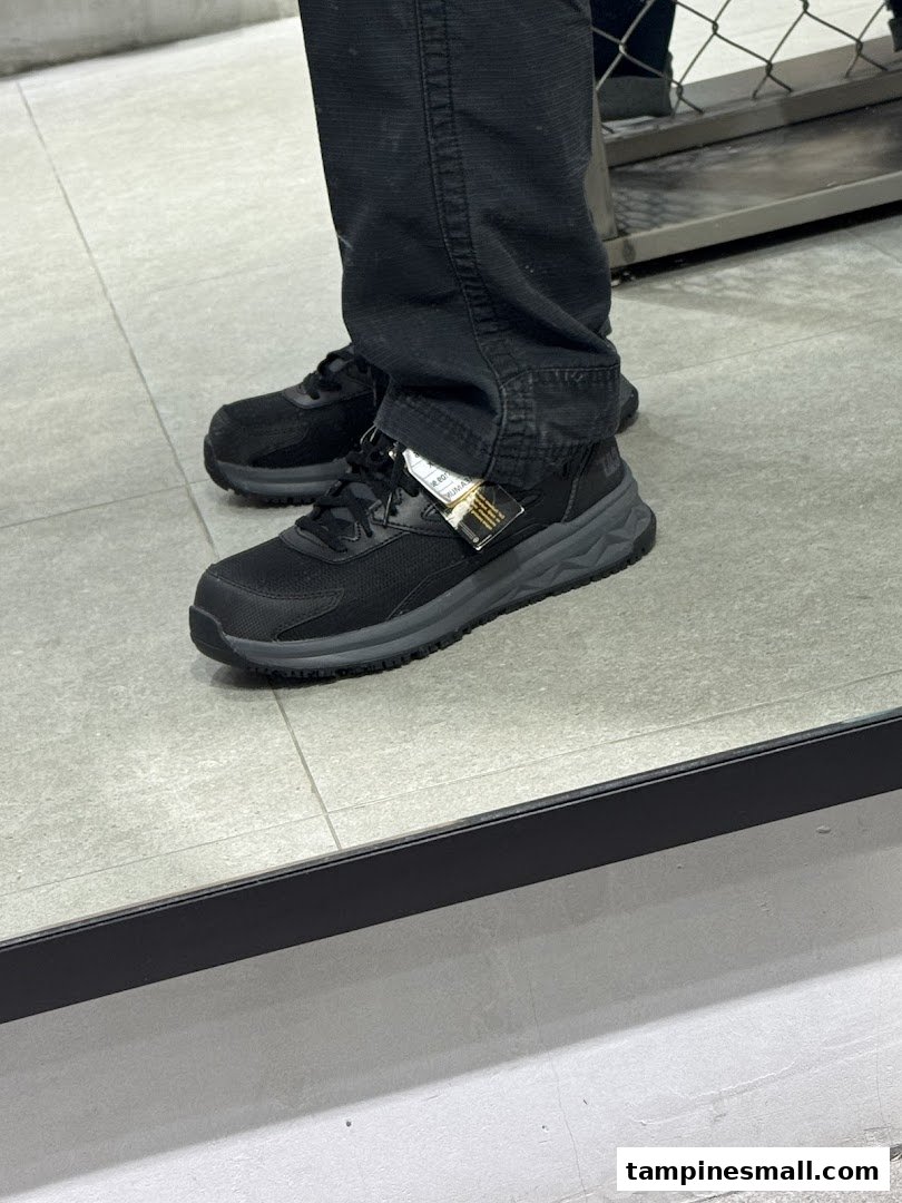 A person trying on a pair of black athletic shoes with a tag visible, reflected in a mirror.