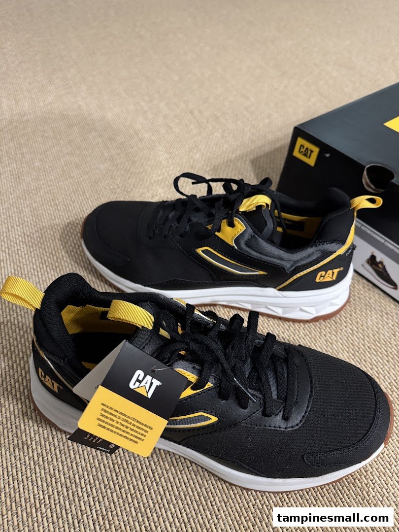 A pair of new black and yellow CAT brand sneakers with their box on a textured surface.