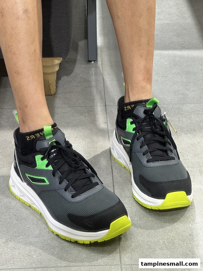 A person's legs wearing a pair of grey and neon green sneakers on a tiled floor.