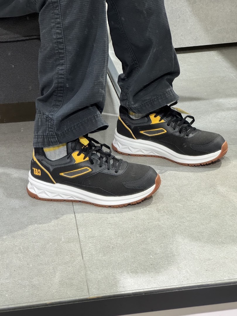 A person wearing black and yellow sneakers with white soles and dark pants, likely trying on shoes.