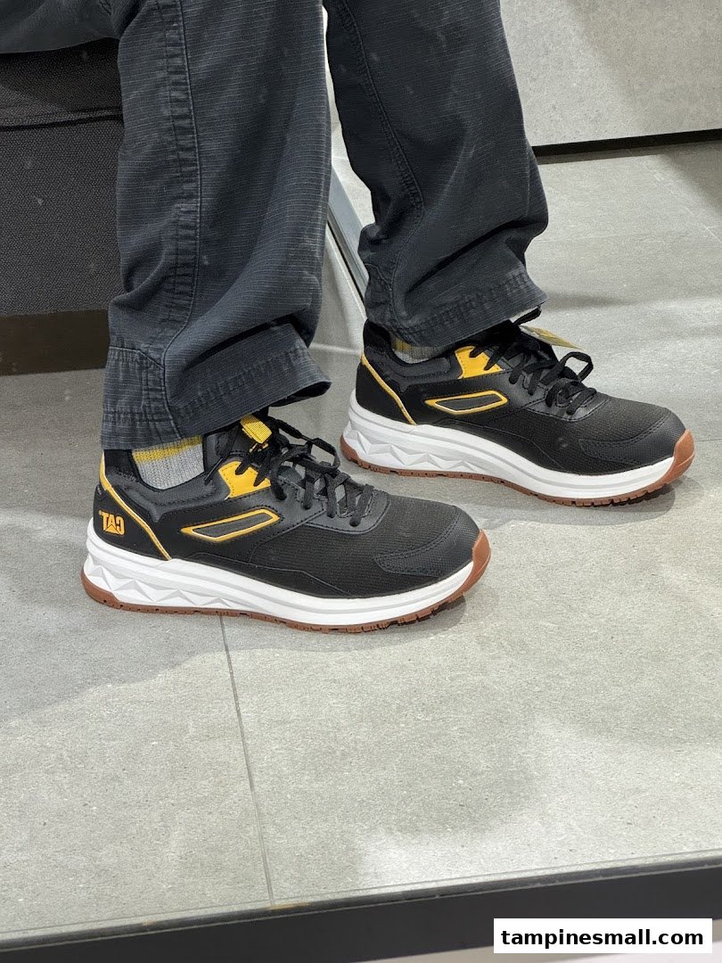 A person wearing black and yellow sneakers with white soles and dark pants, likely trying on shoes.
