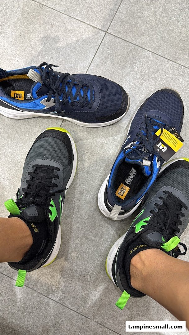 A person trying on a pair of black and neon green sneakers, with another pair of blue sneakers nearby.