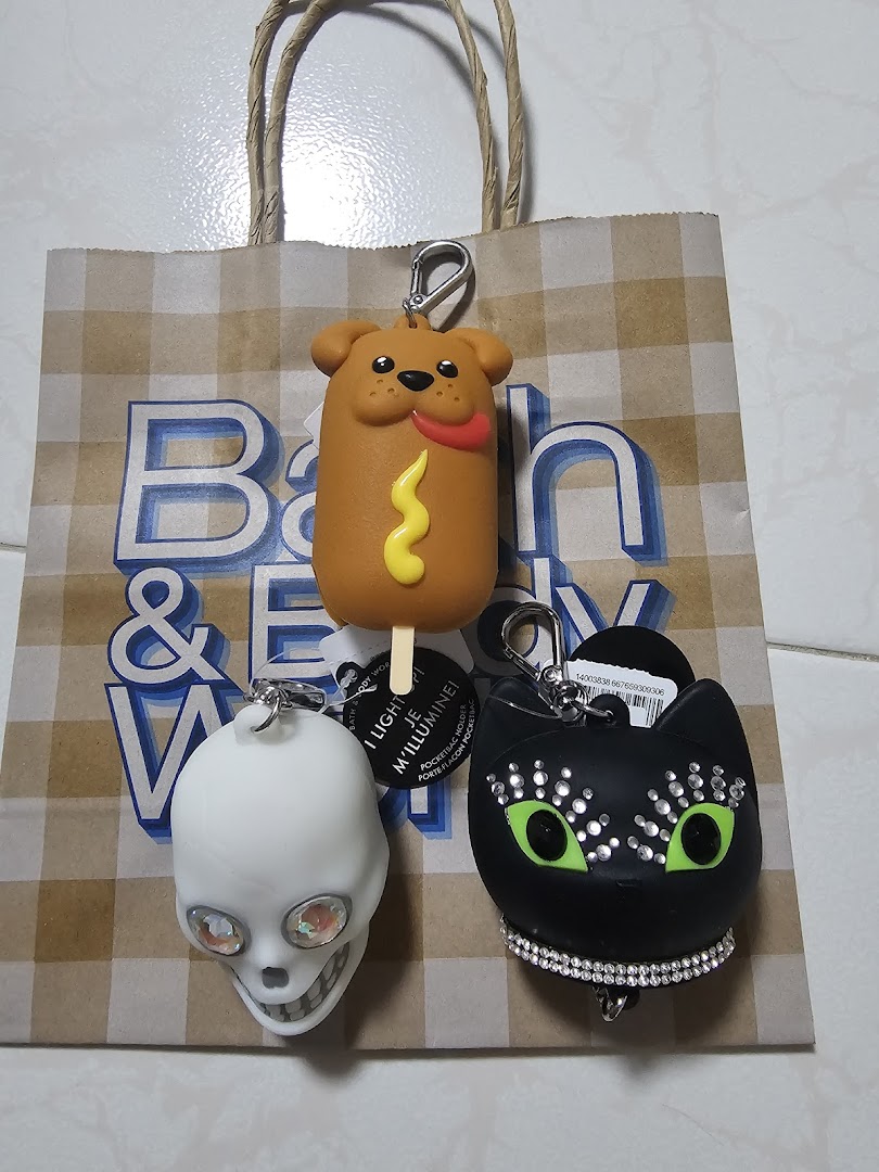Three whimsical Bath & Body Works PocketBac holders, including a hot dog, skull, and black cat, on a branded bag.