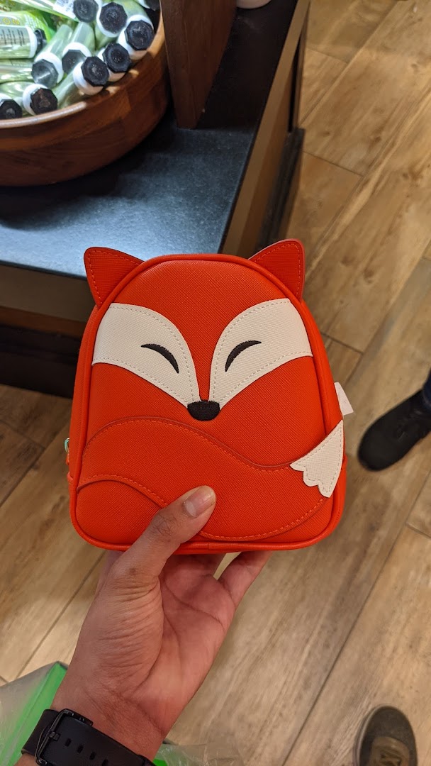A hand holds an orange fox-shaped bag, likely a charming accessory from Bath & Body Works.