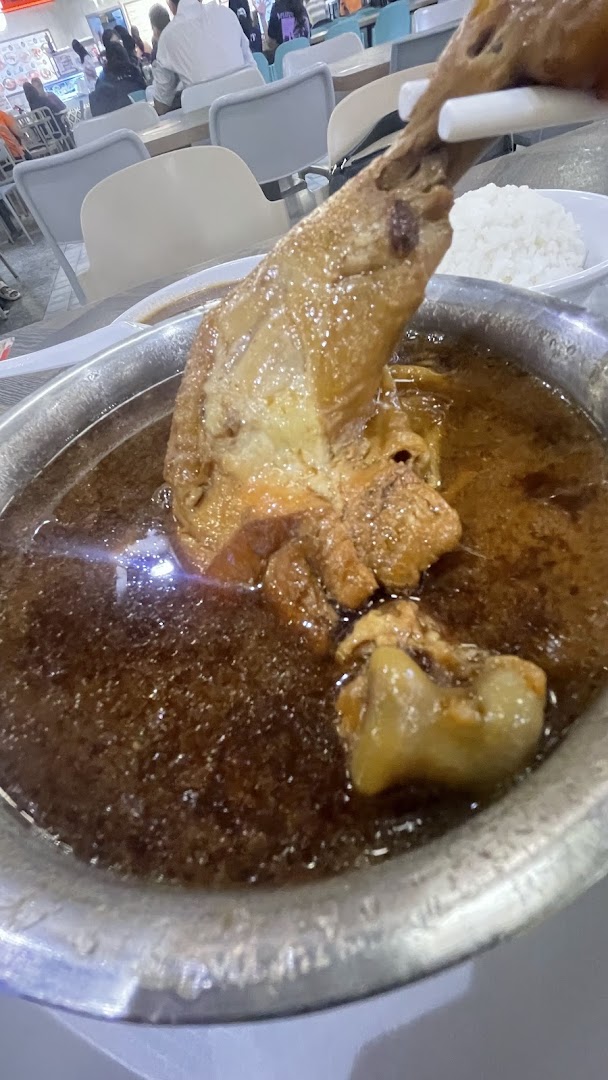 A large braised chicken leg or pork hock in rich dark sauce, served with rice in a food court setting.