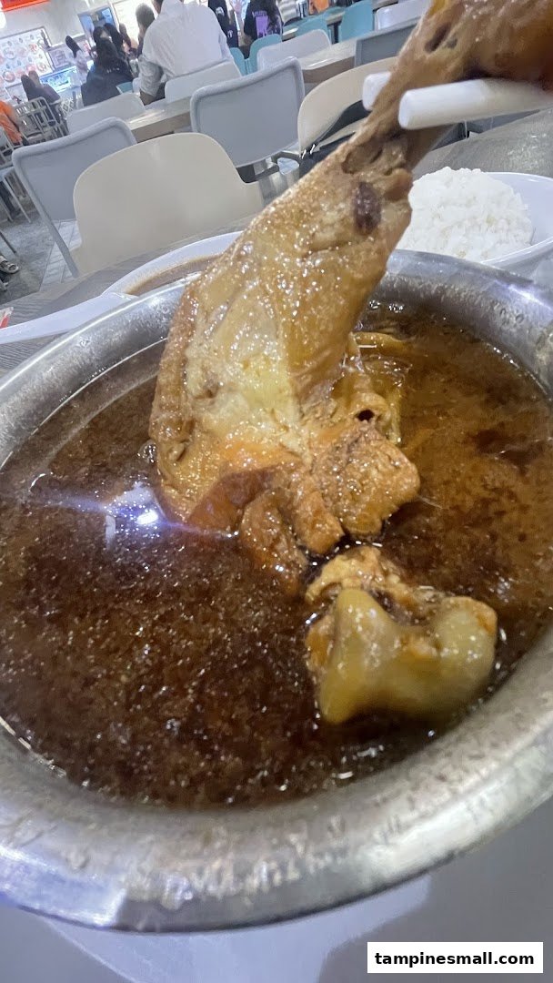 A large braised chicken leg or pork hock in rich dark sauce, served with rice in a food court setting.