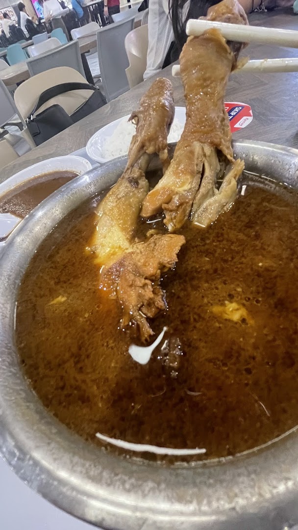 A close-up of braised chicken feet in a rich, dark broth, served in a metal bowl at a food court.