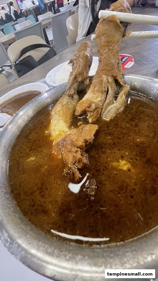 A close-up of braised chicken feet in a rich, dark broth, served in a metal bowl at a food court.
