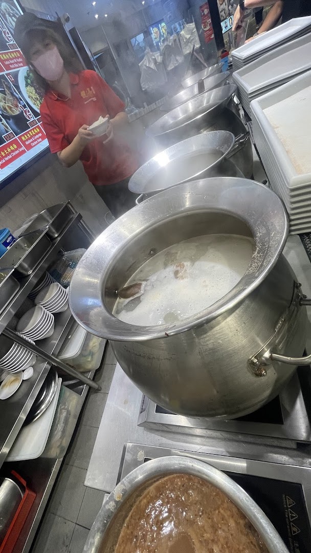 A staff member prepares food in a bustling kitchen with large steaming pots of soup.