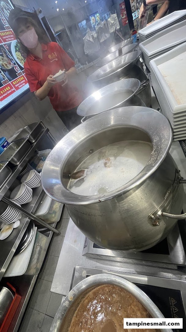 A staff member prepares food in a bustling kitchen with large steaming pots of soup.
