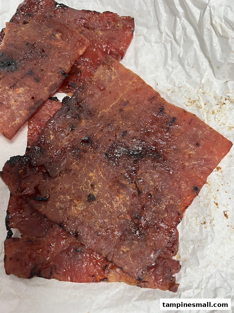 Close-up of several pieces of glistening, grilled bak kwa (barbecued meat jerky) on white paper.