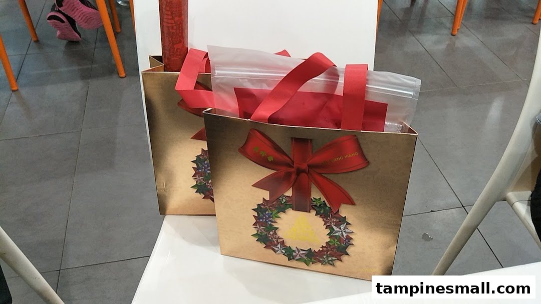 Two festive golden gift bags from Bee Cheng Hiang, adorned with red ribbons and a wreath design.