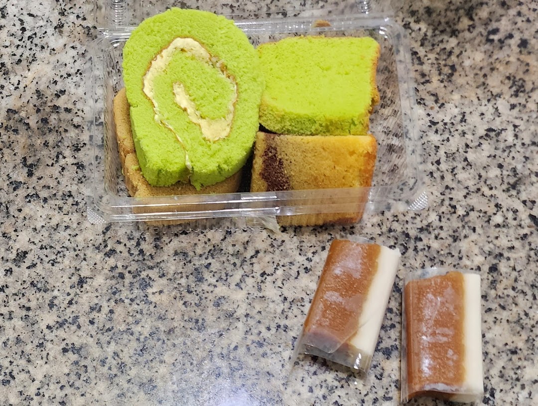 A clear container holds a vibrant green pandan roll cake and other sliced cakes, alongside two wrapped traditional sweets.