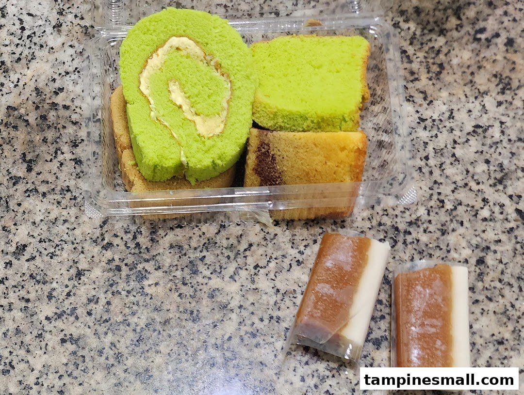 A clear container holds a vibrant green pandan roll cake and other sliced cakes, alongside two wrapped traditional sweets.