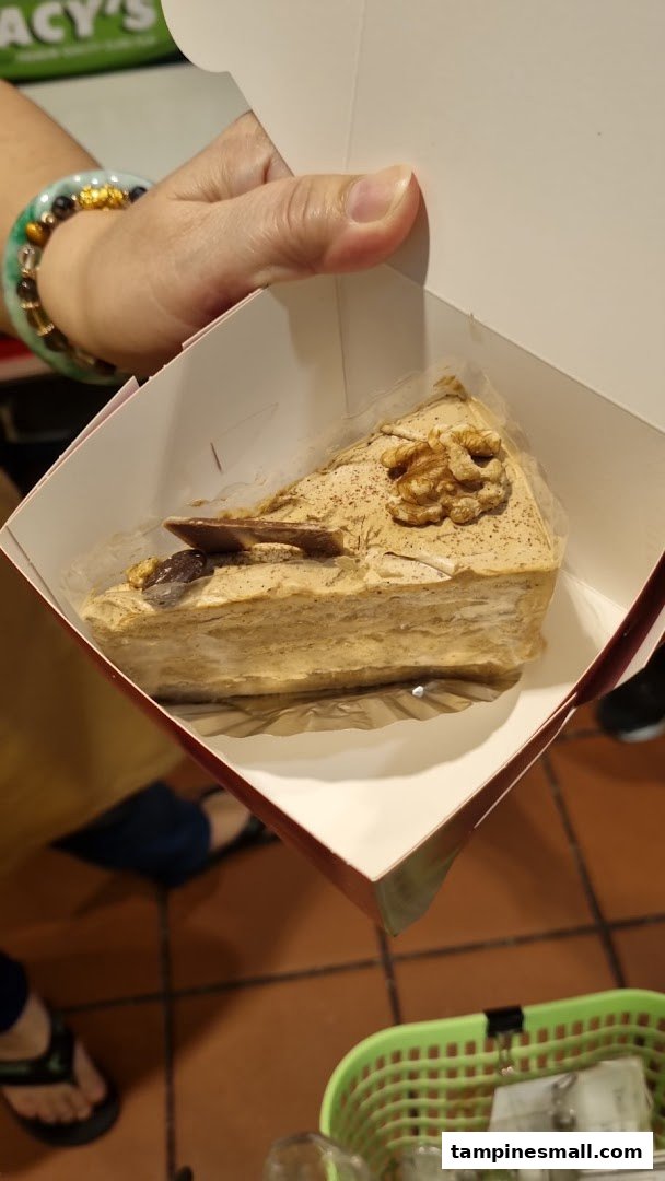 A hand holds a slice of cake in a white takeaway box, featuring walnuts and chocolate.