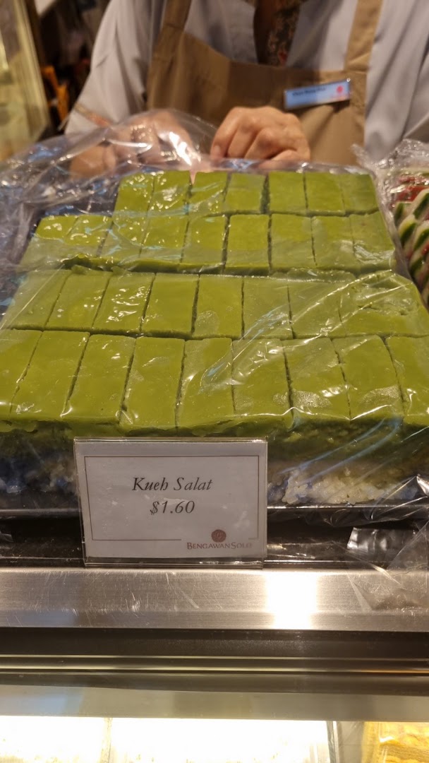 A display of green Kueh Salat pastries with a price tag, behind a counter with a staff member.