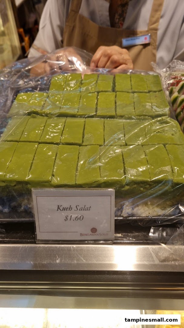 A display of green Kueh Salat pastries with a price tag, behind a counter with a staff member.