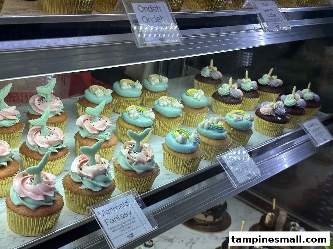 A vibrant display of beautifully decorated cupcakes, including Mermaid Fantasy and Unicorn designs, in a bakery showcase.