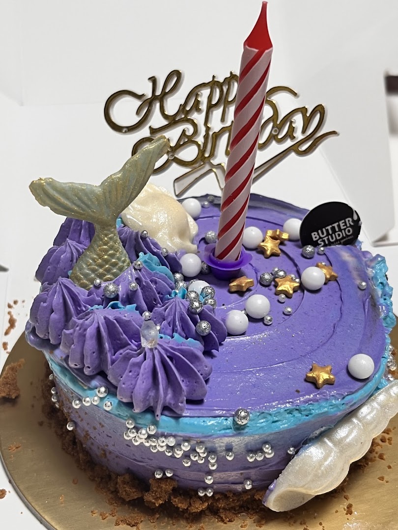 A vibrant purple mermaid-themed birthday cake with a 'Happy Birthday' topper and candle.