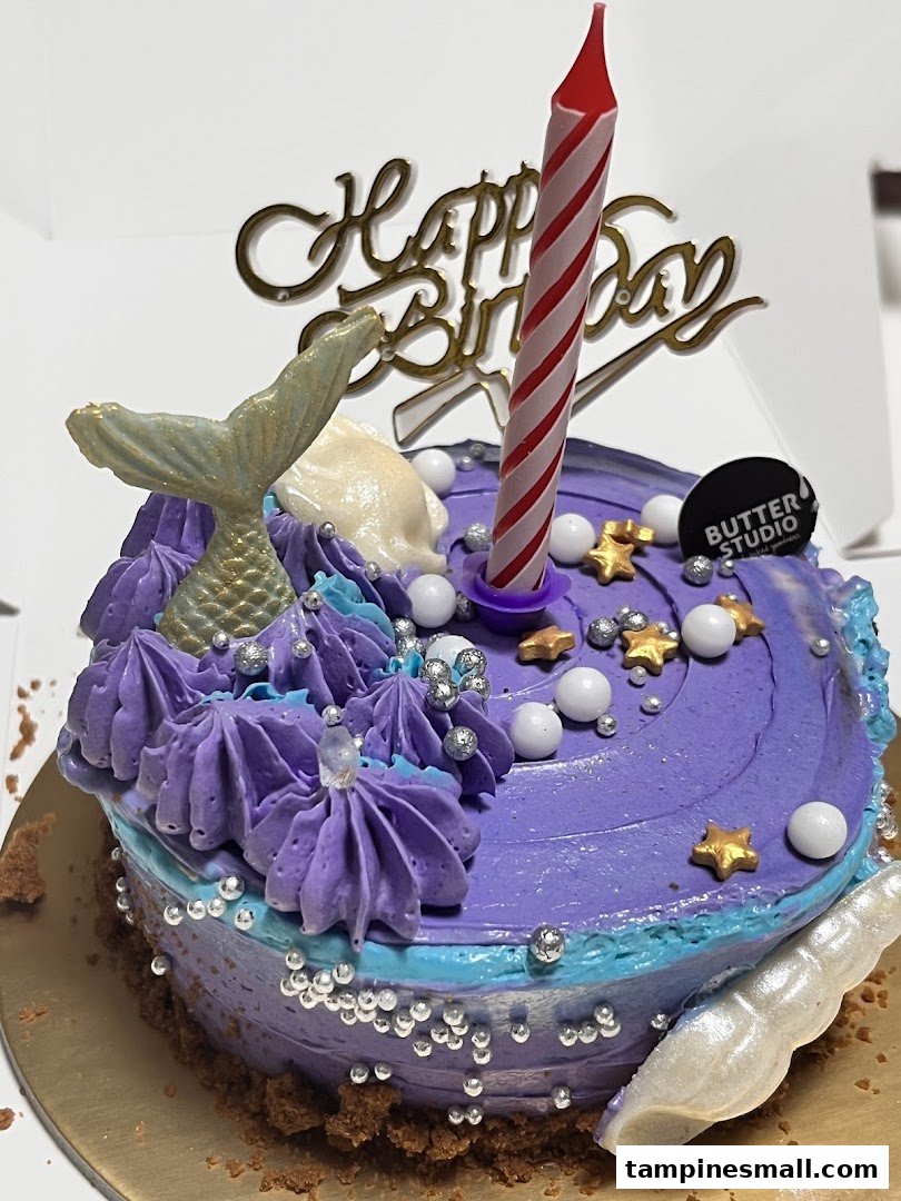 A vibrant purple mermaid-themed birthday cake with a 'Happy Birthday' topper and candle.