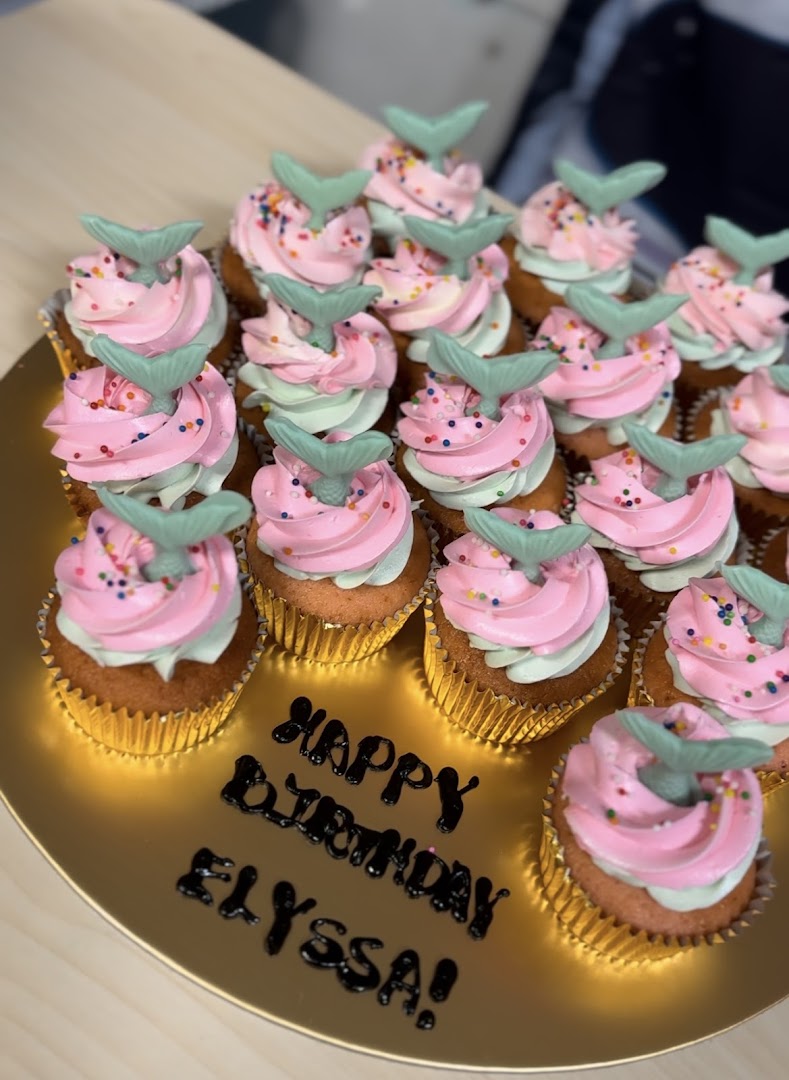 A golden platter filled with pink and mint green mermaid-themed cupcakes with sprinkles and a 'Happy Birthday Elyssa!' message.