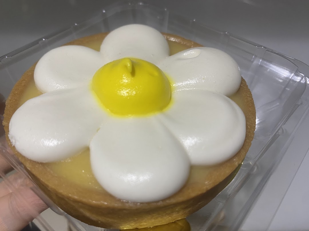 A delightful daisy-shaped tart with white petals and a yellow center, presented in a clear plastic container.