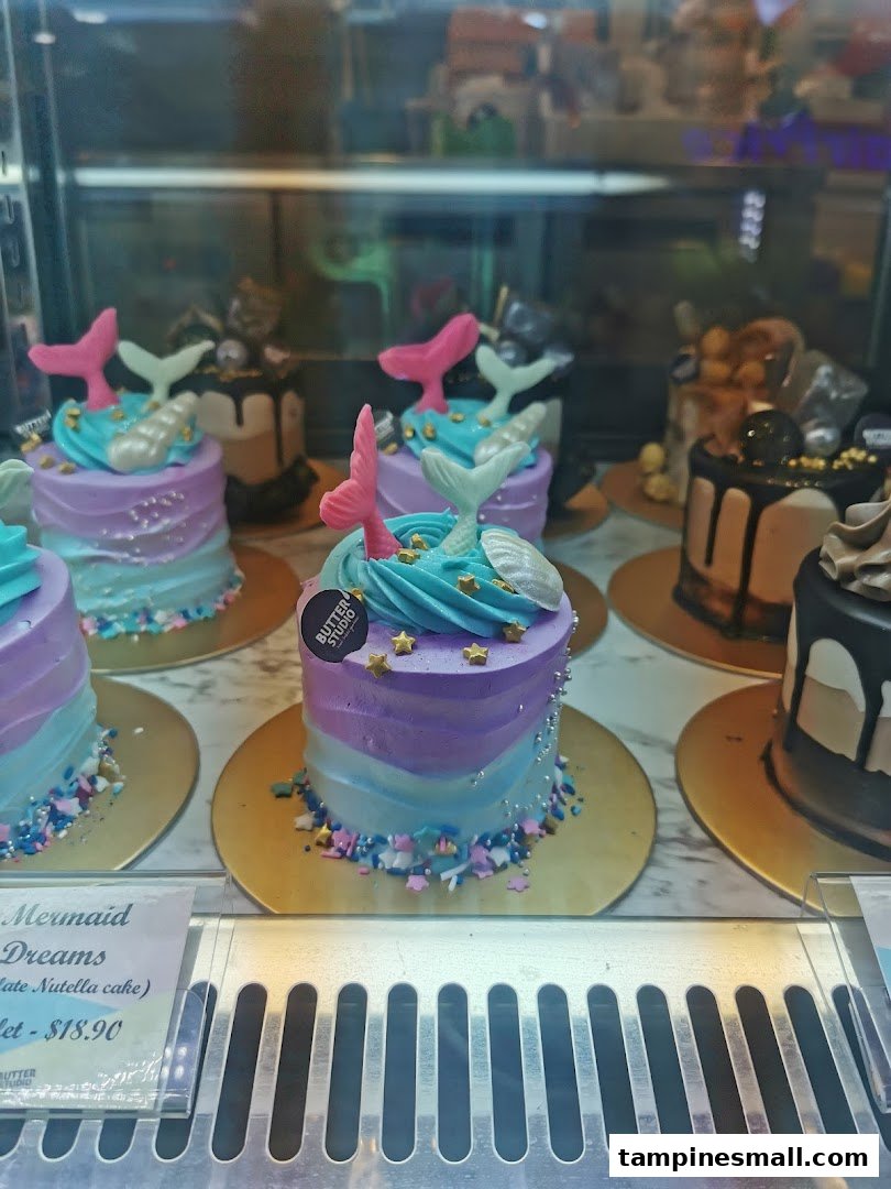 A display of beautifully decorated mini cakes, including 'Mermaid Dreams', in a glass showcase.