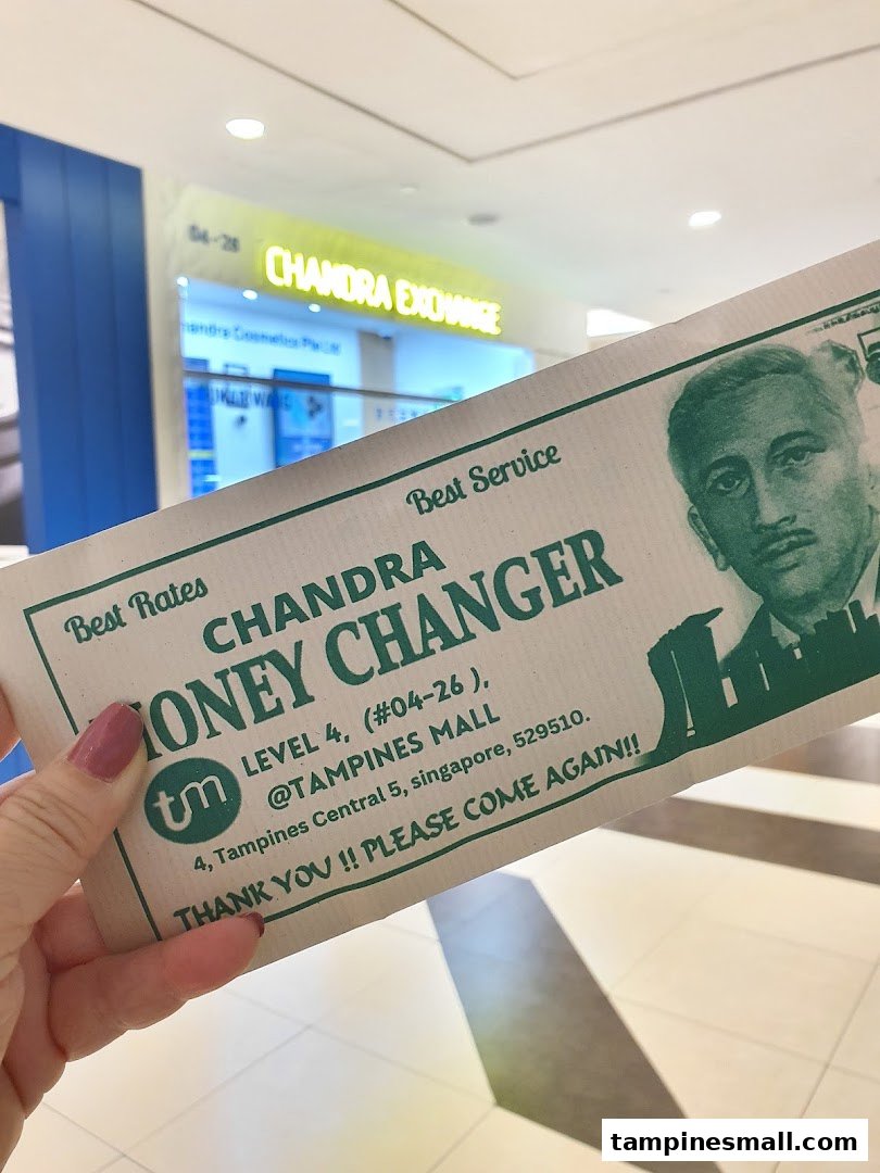 A hand holds a promotional flyer for Chandra Money Changer with the shop's exterior in the background.