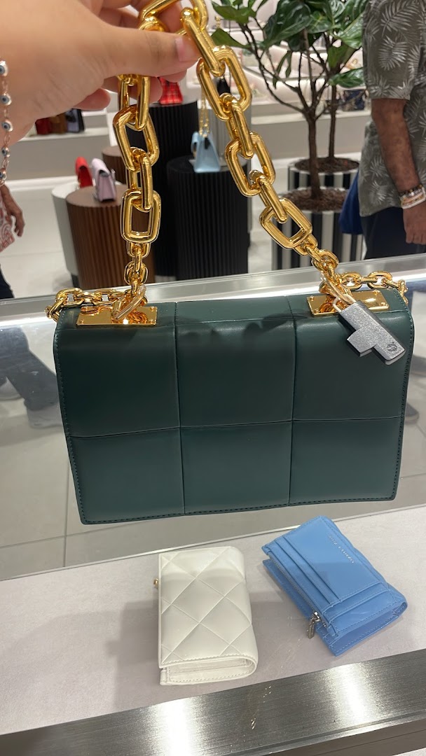 A person holds a dark green quilted handbag with a gold chain, alongside wallets on display at CHARLES & KEITH.