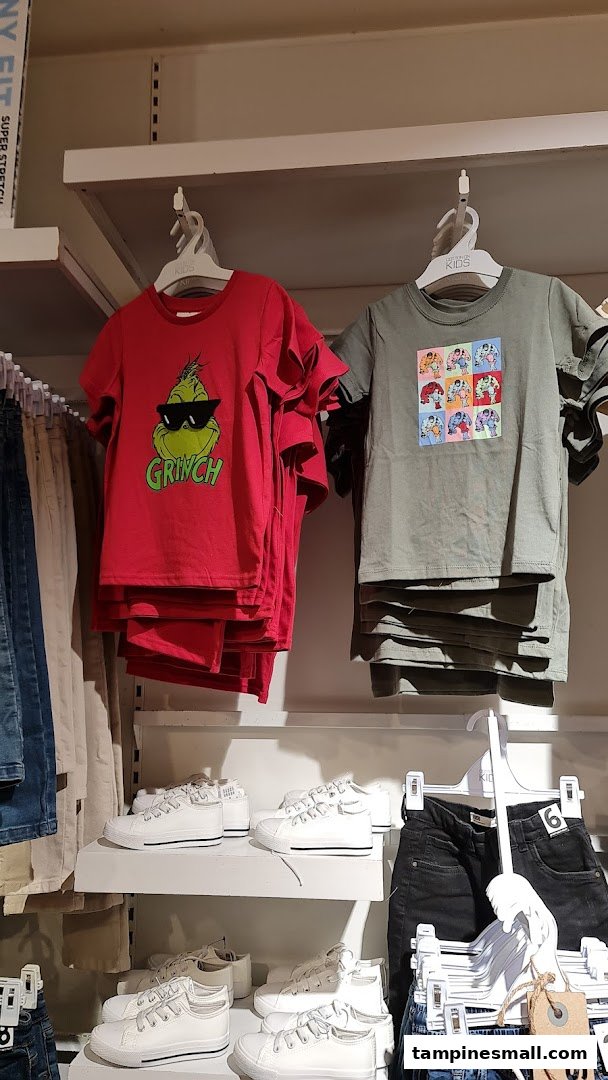 A retail display featuring children's graphic t-shirts (Grinch, pop art) and white sneakers on shelves.