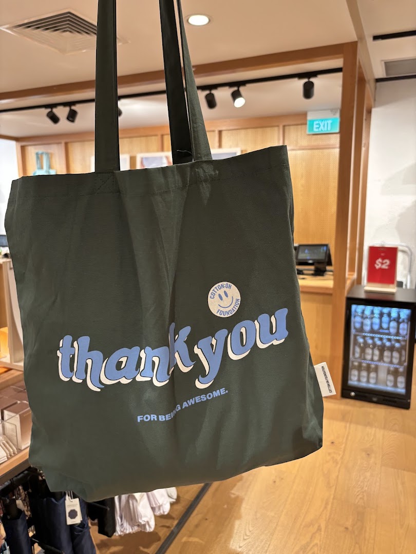 A dark green Cotton On Foundation tote bag with 'thank you' text, displayed inside the store.