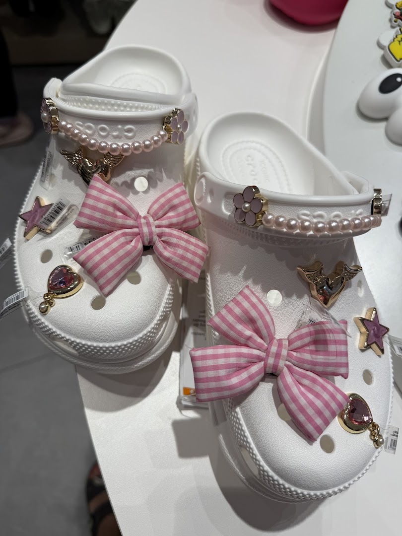 A pair of white Crocs clogs customized with pink bows, pearl chains, and various decorative charms.