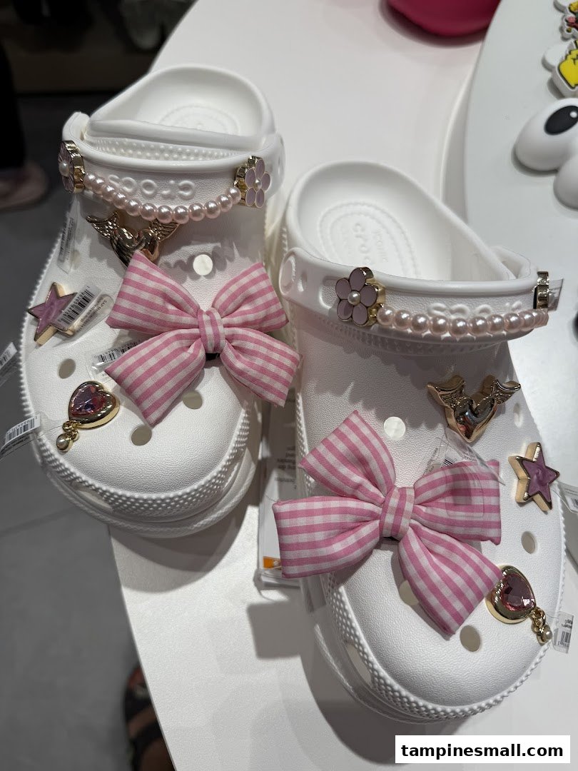 A pair of white Crocs clogs customized with pink bows, pearl chains, and various decorative charms.