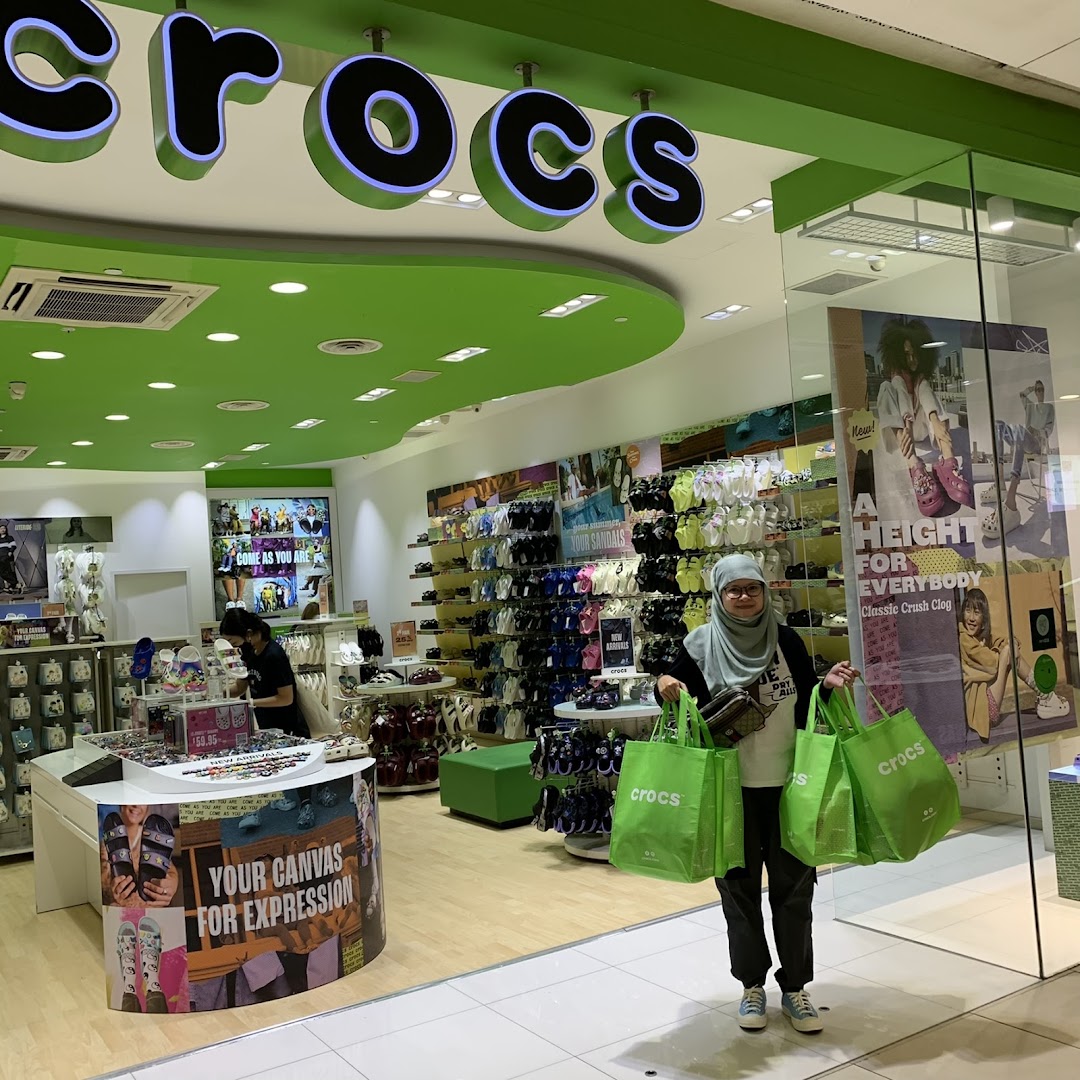 A customer stands outside the Crocs store entrance, holding several bright green shopping bags.