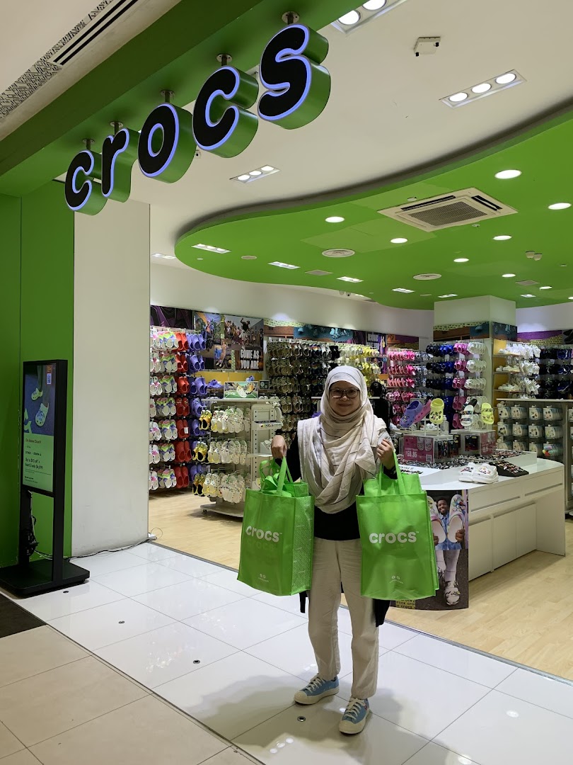 A shopper stands outside the vibrant Crocs store entrance, holding two green shopping bags.