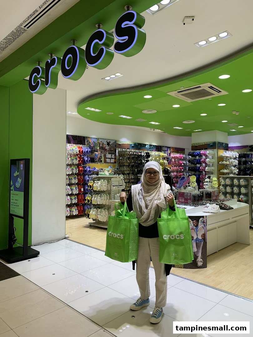 A shopper stands outside the vibrant Crocs store entrance, holding two green shopping bags.