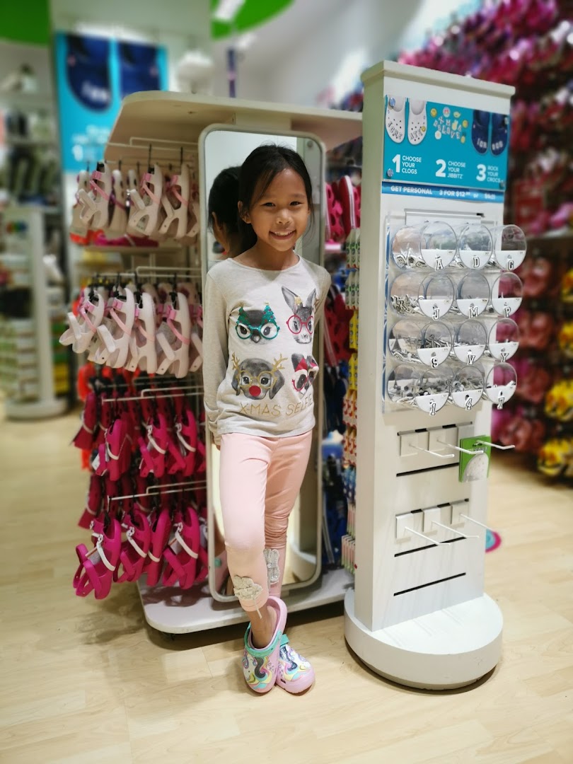 A smiling young girl stands in a vibrant Crocs store, surrounded by colorful footwear and Jibbitz displays.