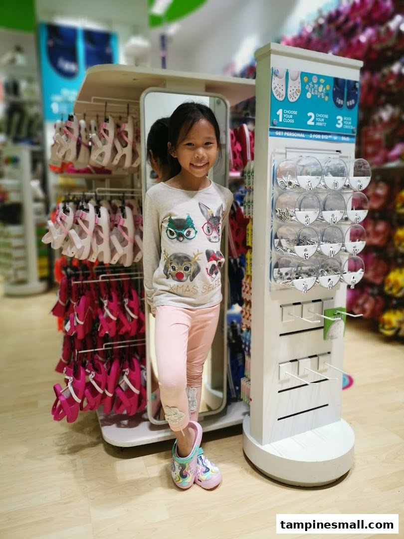 A smiling young girl stands in a vibrant Crocs store, surrounded by colorful footwear and Jibbitz displays.