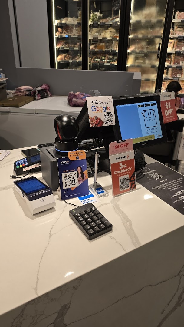 A well-lit checkout counter at Cut Butchery, featuring payment terminals and refrigerated meat displays in the background.