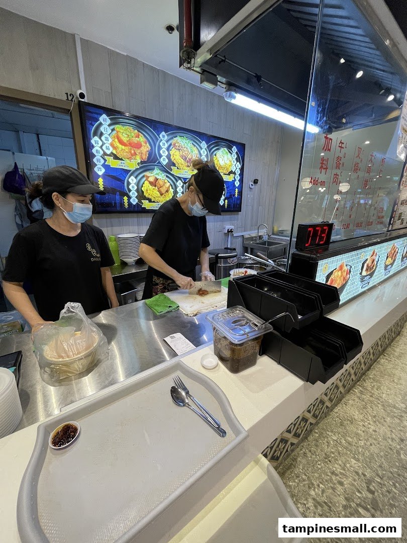 Two staff members preparing food at a counter with a digital menu board displaying scrambled egg rice dishes.