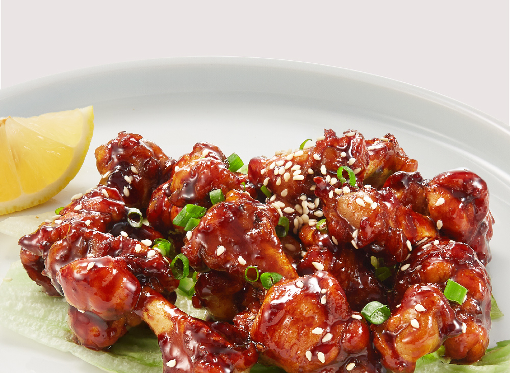Close-up of glazed chicken pieces garnished with sesame seeds, green onions, and a lemon wedge.
